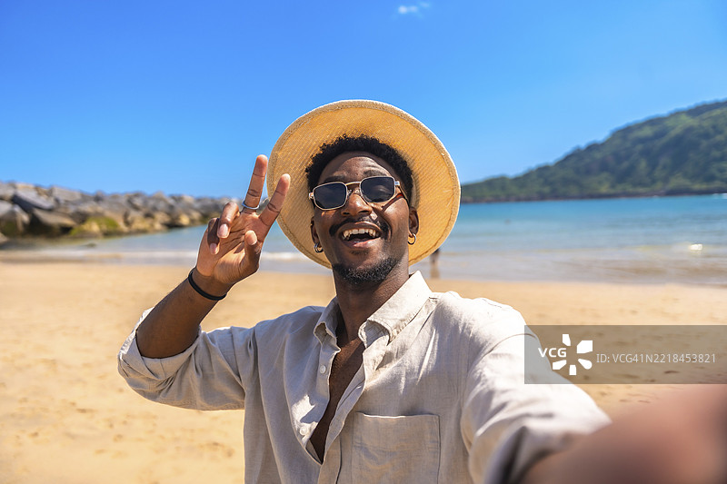 年轻男子戴着帽子和太阳镜，在热带海滩上比出胜利手势自拍图片素材