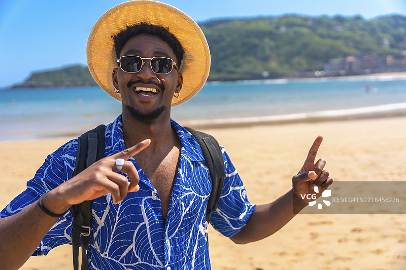 快乐的黑人游客在美丽的海滩上微笑着，双手食指指向天空，享受阳光和海洋。图片素材