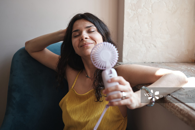 夏季炎热，生活在没有空调的环境中。年轻女性坐在家中窗边，手持便携式电动迷你风扇，享受炎热天气中的清新空气，感到凉爽。图片素材