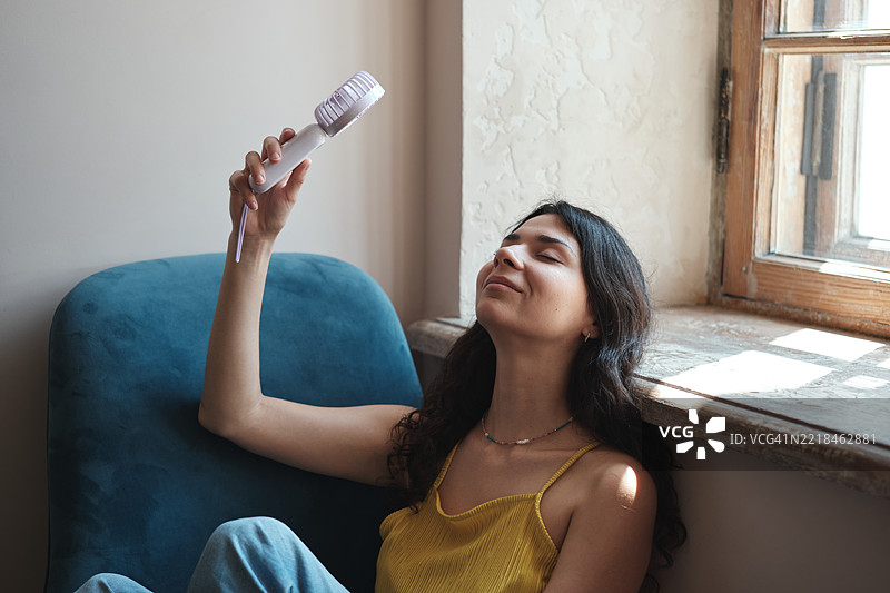 年轻女性在家中忍受夏季炎热，使用便携式电动迷你风扇，享受清新空气，在热浪中降温。图片素材