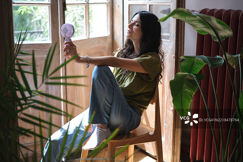 年轻女性在窗边使用便携式电动迷你风扇，享受新鲜空气，在家中降温，忍受夏季的炎热。图片素材