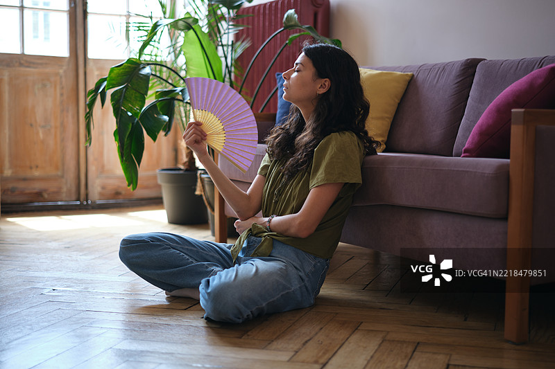 夏季热浪。年轻女性在家中忍受夏季炎热，手持扇子，在没有空调的公寓里降温。图片素材