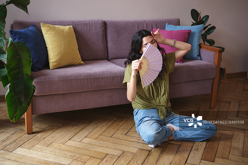 夏季热浪。年轻女性在家中感受夏季炎热，坐在地板上手持扇子，在没有空调的公寓里降温。图片素材