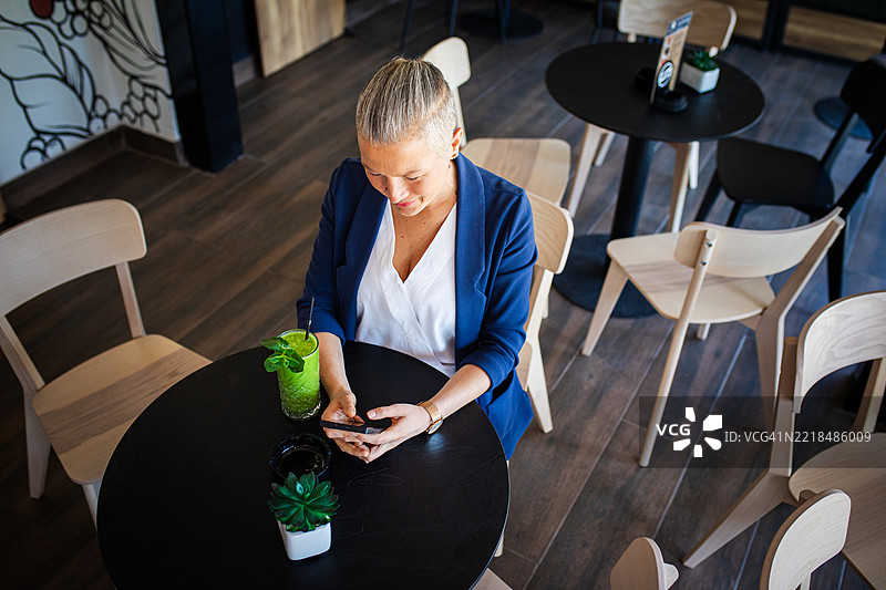 快乐的女性在咖啡店里使用手机，手边有一杯绿色果昔图片素材