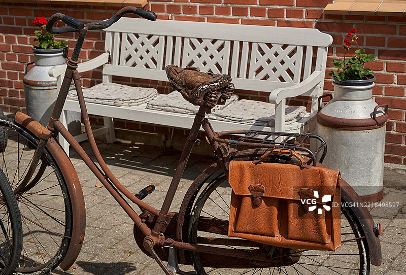 一辆非常旧的生锈自行车，坐垫破损，车架上放着一个旧皮公文包，停在一个舒适的长椅前，长椅旁边用牛奶罐做花盆。图片素材