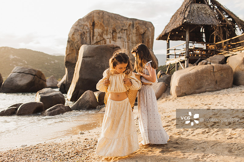 两个年轻女孩在沙滩上走，周围是乡村小屋和岩石图片素材