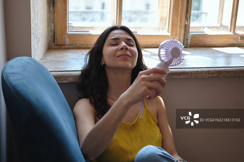 年轻满意的女性坐在窗边，使用便携式电风扇，享受新鲜空气，在家中降温，抵御夏季热浪。图片素材