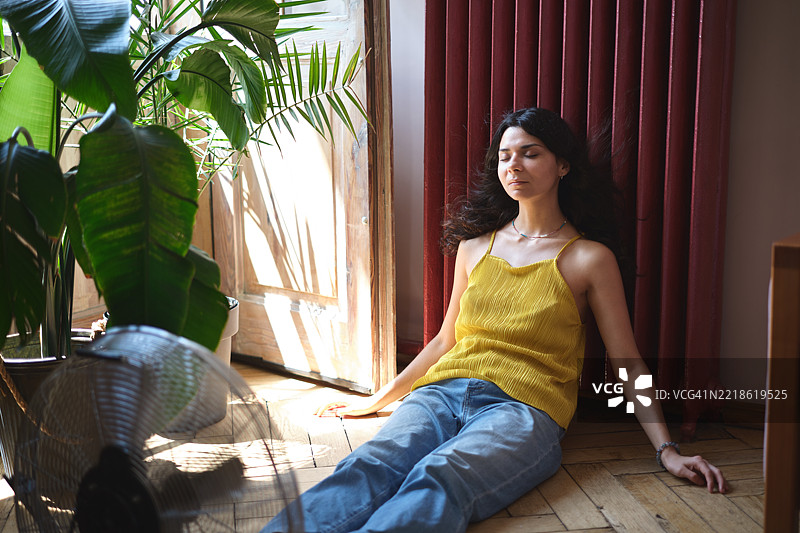 在没有空调的情况下保持凉爽。夏季炎热。年轻的放松女性坐在家中地板上，靠近电风扇，在热浪中降温。图片素材