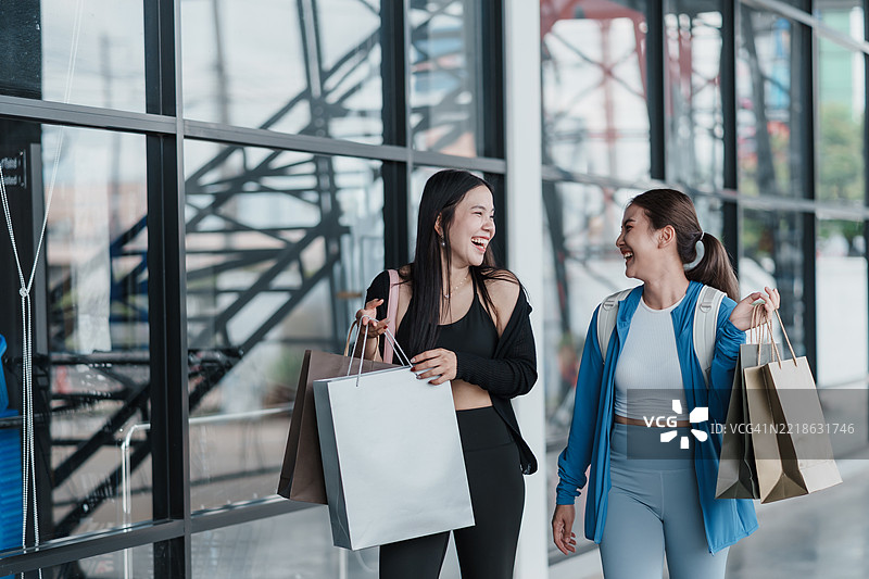 亚洲女性一边走一边笑，手里提着购物袋。友谊生活方式，零售购物，健身时尚，运动服购买，现代消费体验，快乐时刻。图片素材