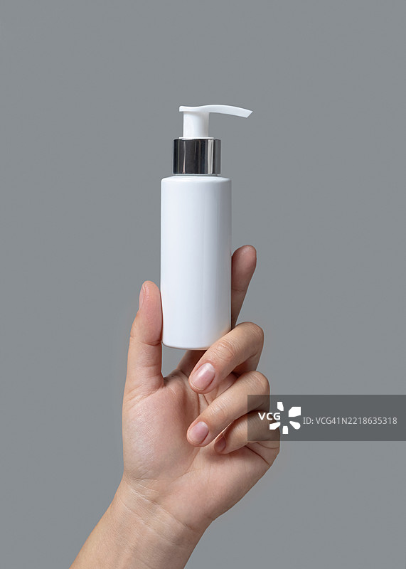 女性手握白色化妆品泵瓶，背景为灰色特写，模版，留白空间图片素材