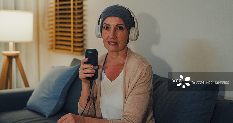 戴着耳机的老年女性癌症患者，头上裹着头巾，在家中客厅的沙发上分享她的癌症康复故事，夜晚的氛围。与癌症共存的概念。图片素材