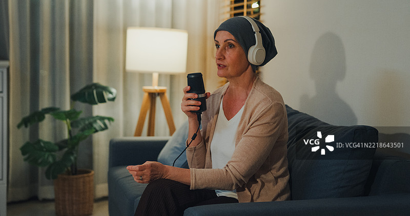 戴着头巾的老年女性癌症患者，佩戴耳机，通过播客分享她的癌症康复故事，坐在家中客厅的沙发上，夜晚的氛围。与癌症共存的概念。图片素材
