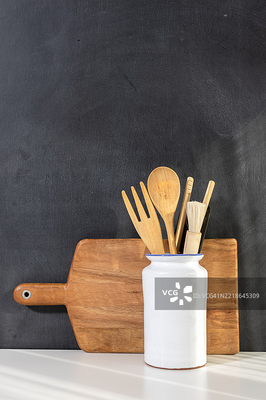 白色陶瓷容器中的木质厨房用具和靠在黑板墙上的木砧板，留有空白空间，营造出温馨的厨房场景图片素材