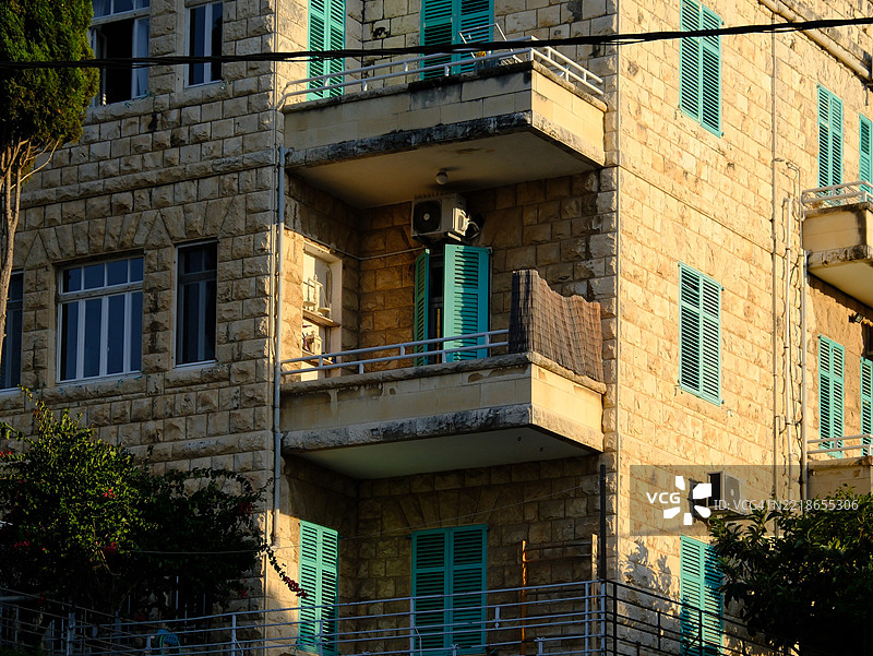 以色列海法一座石头建筑的美丽照片，配有绿松石色的百叶窗和阳台。阳光的金色光芒照亮了石材的纹理，突显了建筑细节和绿意。图片素材