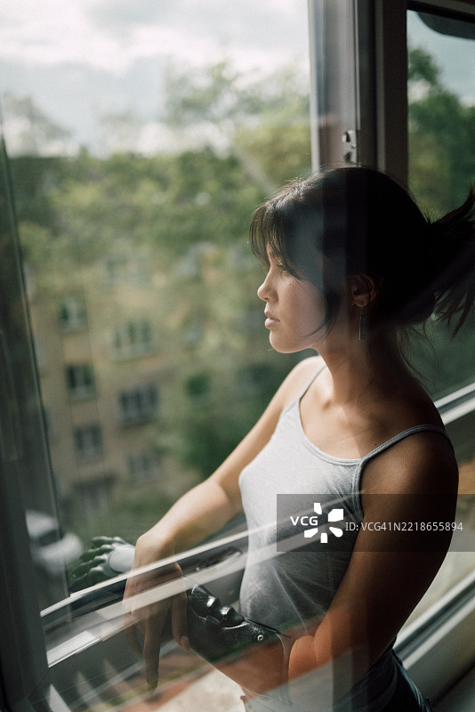 阳光下，年轻女性站在开着的窗边，深思熟虑，手臂为仿生手臂。图片素材