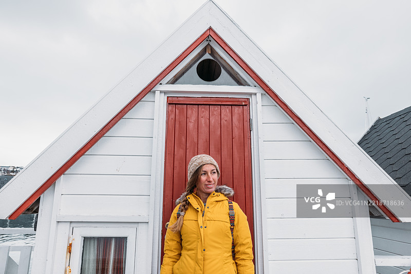 穿着黄色外套的微笑女性站在温馨小屋的红色门旁图片素材