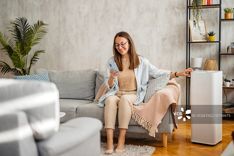 年轻女性在家沙发上用手机开启空气净化器图片素材