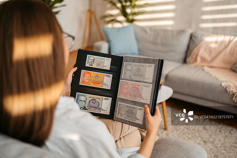 年轻女性坐在客厅沙发上，查看一册旧钞票收藏。图片素材