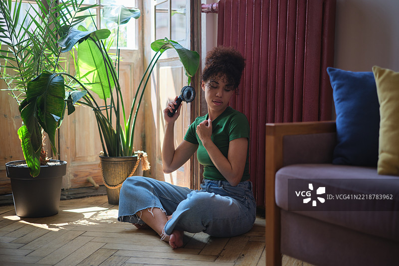 夏季炎热。非裔美国年轻女孩在家中使用便携风扇，因中暑而感到不适，坐在窗边享受新鲜空气，在室内降温度过热浪。图片素材