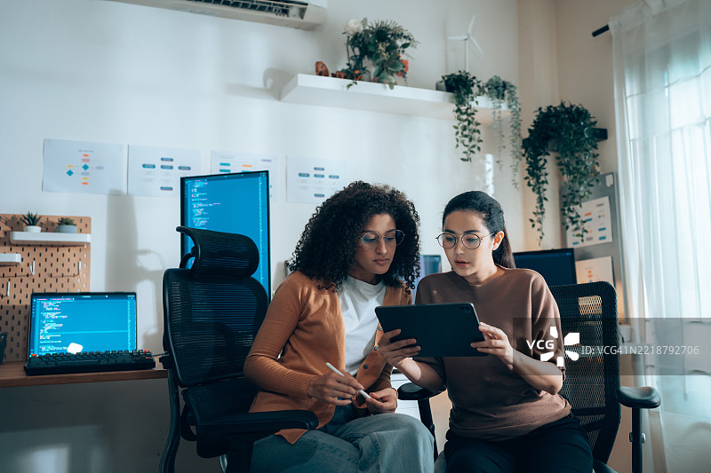 女性科技工作者在平板电脑上合作进行用户界面审查，背景中有编码显示器。图片素材