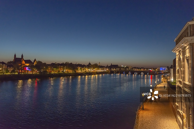 荷兰马斯特里赫特，夜晚的马斯河景观，灯光照亮的桥梁和滨水步道，天空晴朗。图片素材