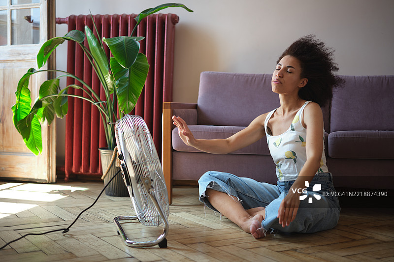 快乐满足的非裔美国人卷发女性在家中使用电风扇，享受炎热夏日的清新空气。图片素材