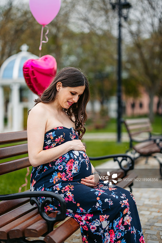 中年孕妇坐在公园长椅上，背景中有粉色气球。图片素材