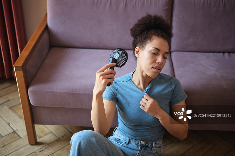 夏季热浪。特写镜头下，一位疲惫的非裔美国女性手持便携式迷你风扇，因酷热而感到痛苦，在家中享受清新空气，降温。图片素材