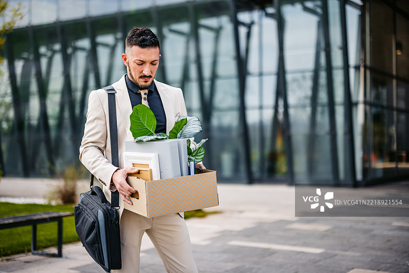 悲伤的中年商人手里拿着办公桌上的物品，站在一座企业大楼前。图片素材