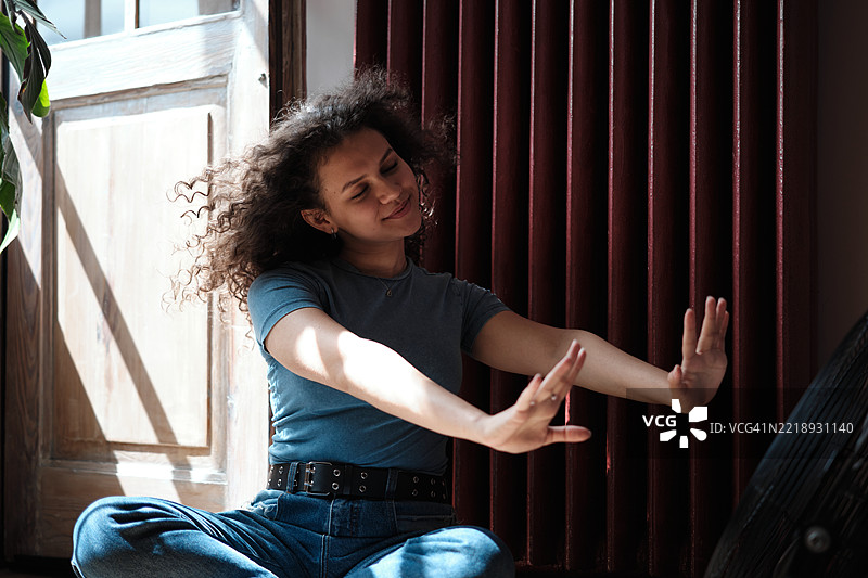 满足的卷发Z世代女孩坐在电风扇旁，享受炎热夏日的清新空气，女性在热浪中生存。图片素材