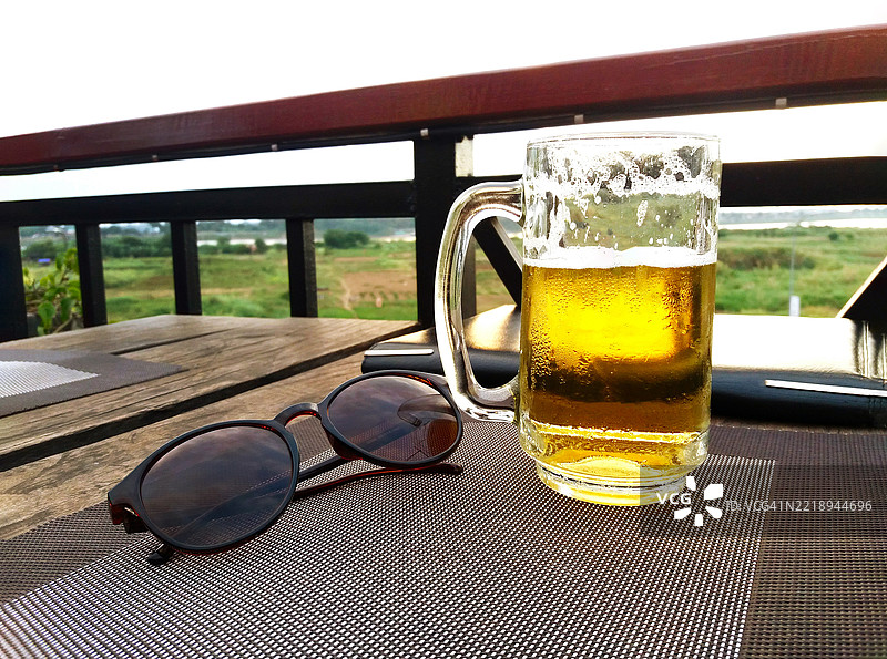 桌子上放着一杯啤酒和一副棕色太阳镜，背景是模糊的天空和绿色树木，留有空白空间。图片素材