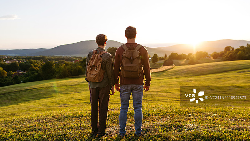父亲和青少年儿子背对背一起徒步旅行，欣赏日落。图片素材