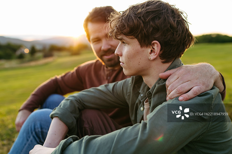 父亲和青少年儿子坐在山顶，欣赏徒步旅行后的自然风光。图片素材