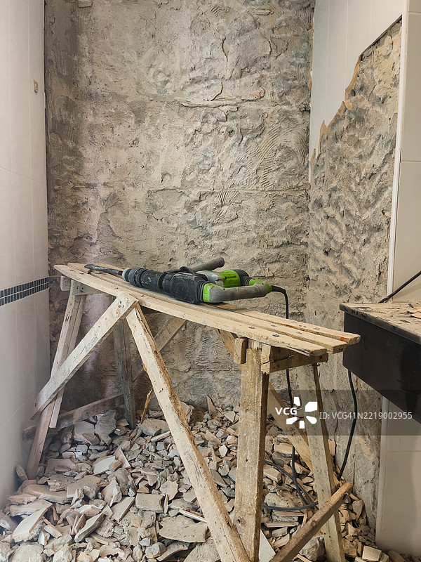 部分拆除的浴室墙壁，地上有破碎的瓷砖和杂物，木制脚手架平台上放着电动工具。图片素材