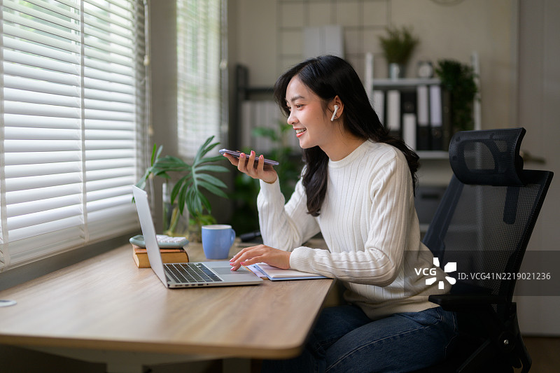 年轻的亚洲女性在家工作，使用笔记本电脑发送语音信息，微笑着享受高效的办公日。图片素材