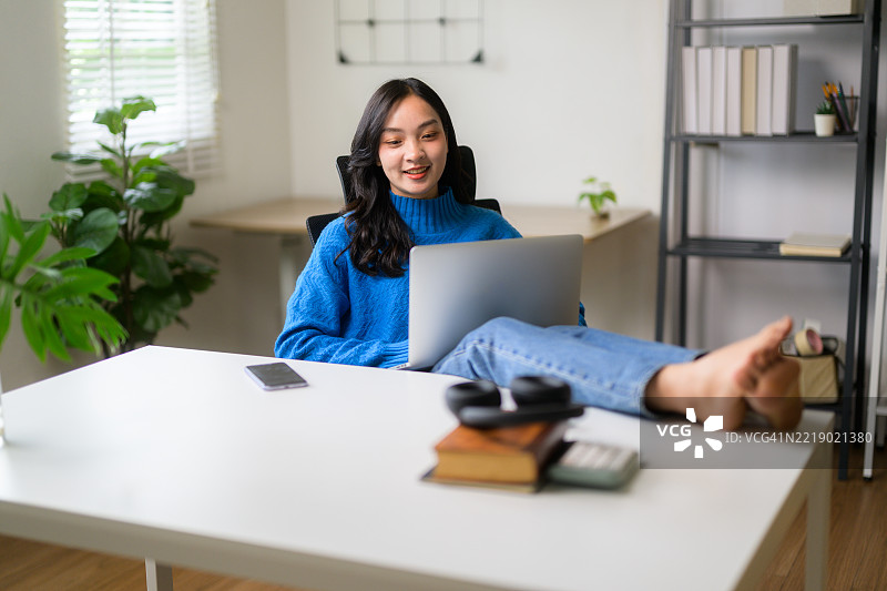 年轻的亚洲女性在家工作，舒适地坐在桌子旁，双脚翘起，使用笔记本电脑。图片素材