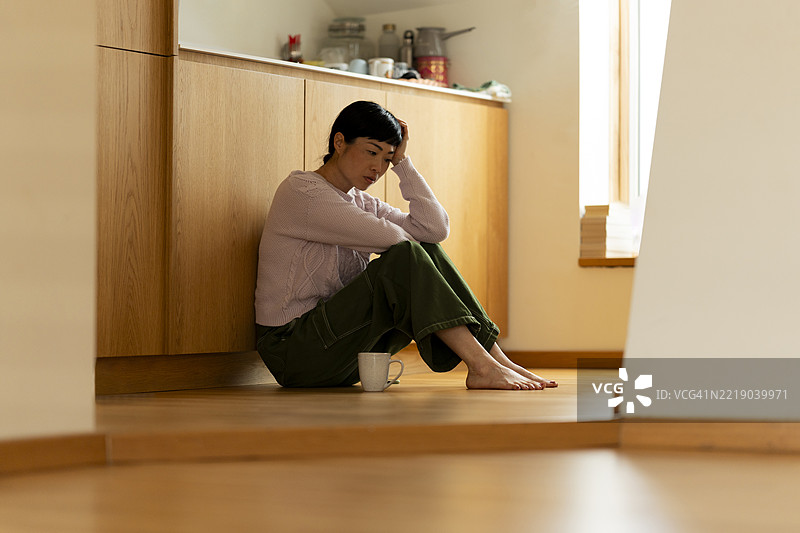 伤心的亚洲女性坐在厨房的地板上，感到孤独。图片素材