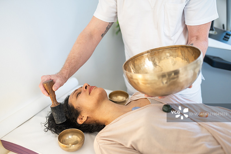 中年男性治疗师在室内使用唱碗为一位西班牙裔女性进行声音疗法图片素材