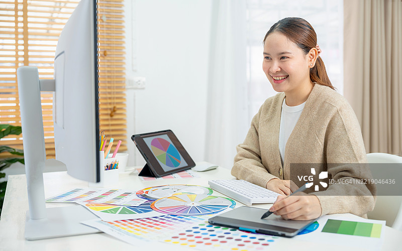亚洲年轻女性平面设计师在现代数字工作室使用数字平板和数位笔进行图形设计。她在电脑笔记本上选择颜色。图片素材