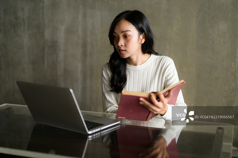 亚洲学生在家中舒适地阅读书籍，同时使用笔记本电脑，全神贯注于在线课程。图片素材