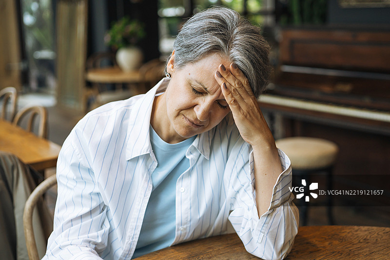 一位头痛的女性坐在咖啡馆的桌子旁，手放在额头上。图片素材