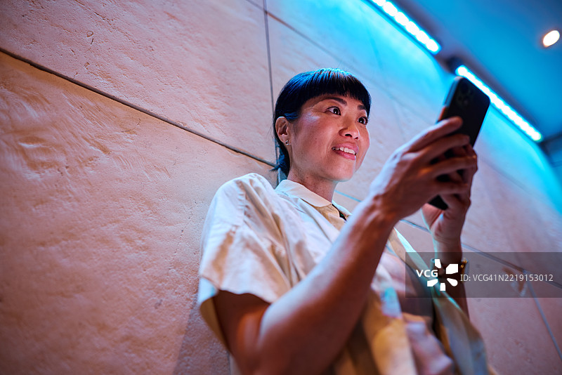 亚洲女商人在现代办公室中使用智能手机，办公室内有蓝色LED灯光。图片素材