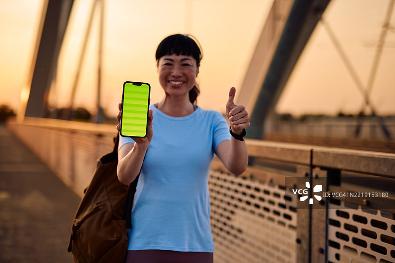 微笑的成熟的女性在日落时分的桥上展示带有绿色屏幕的智能手机，并竖起大拇指。图片素材