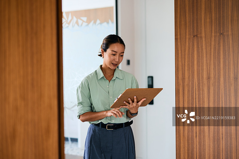 女商人在办公室走廊中边走边查看手中的夹板笔记。图片素材