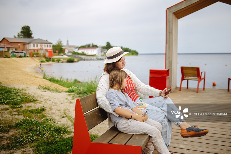 一位加大码妈妈和孩子坐在大湖边的长椅上，两人都在使用智能手机。图片素材