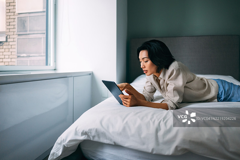 女人在明亮的窗边放松地躺在床上使用平板电脑图片素材