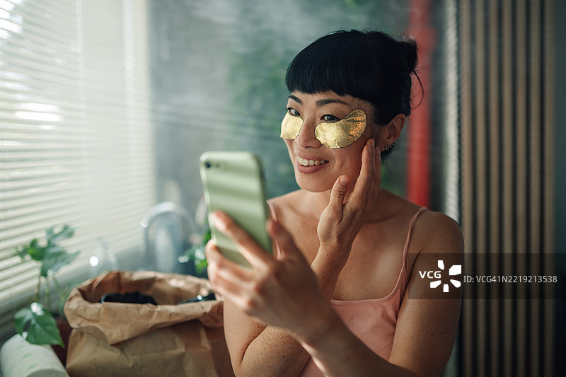 微笑的女性在家中使用智能手机并敷金色眼贴图片素材