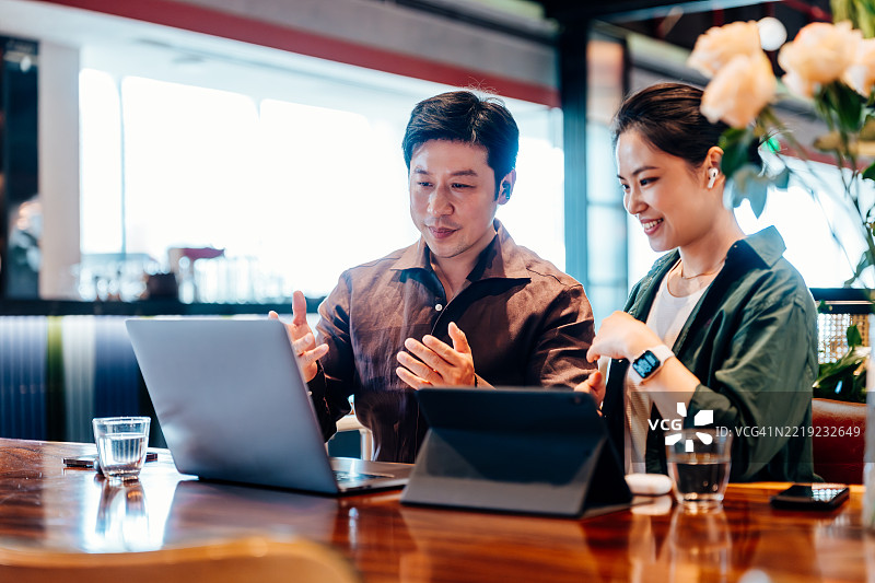 亚洲商务人士在咖啡馆的隔间里戴着耳机开会，使用笔记本电脑共同工作。商务人士总是忙于使用笔记本电脑和手机。图片素材