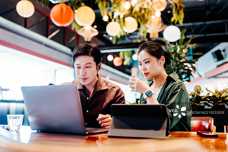 亚洲女商人与男商在咖啡馆的隔间里戴着耳机，使用笔记本电脑开会并共同工作。商界人士总是忙于使用笔记本电脑和手机。图片素材