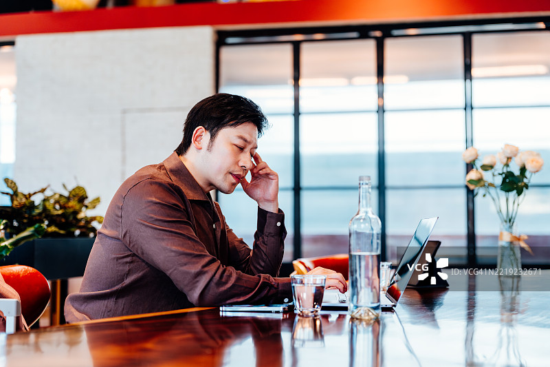 年轻的亚洲商人在咖啡馆里闭着眼睛工作，使用笔记本电脑，展现出严肃和沉思的情感。图片素材